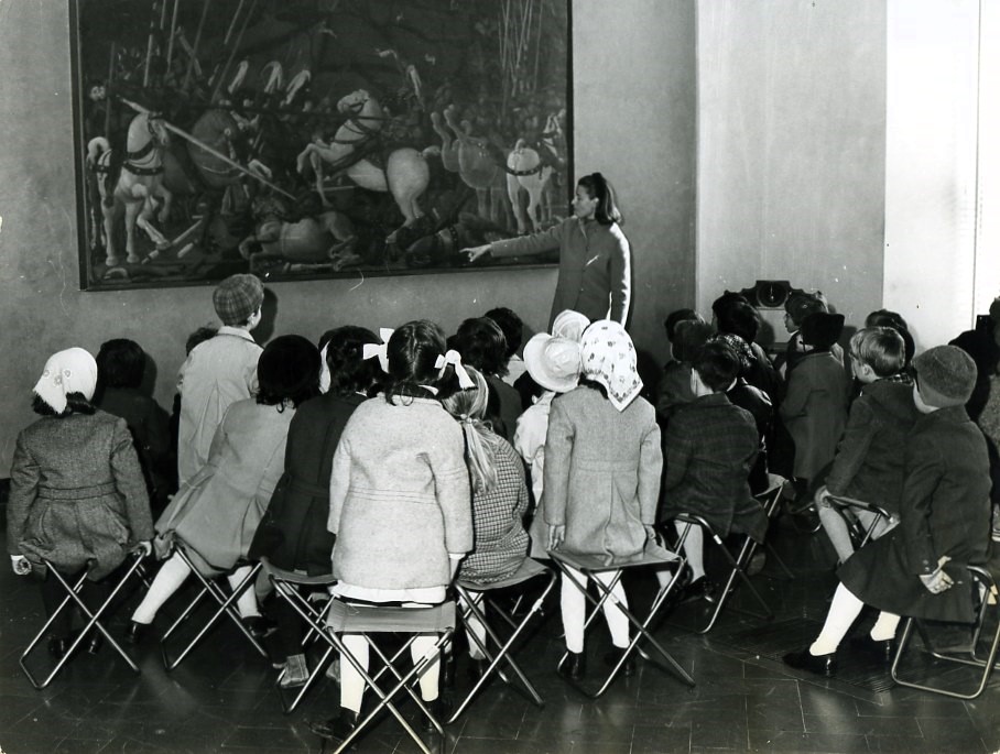 Un gruppo di bambini in abiti invernali è seduto su sgabelli pieghevoli, mentre una guida indica un grande dipinto storico all'interno di una galleria d'arte.