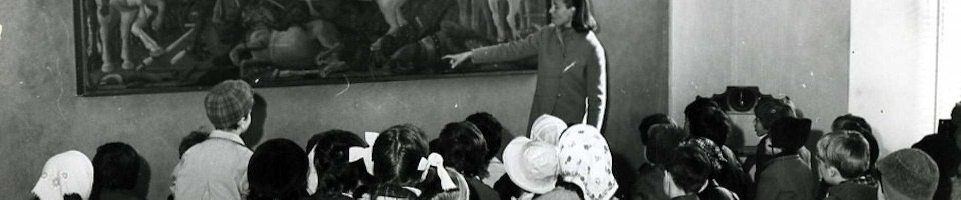 Maria Fossi Todorov, fondatrice Sezione Didattica Uffizi Un gruppo di bambini in abiti invernali è seduto su sgabelli pieghevoli, mentre una guida indica un grande dipinto storico all'interno di una galleria d'arte.