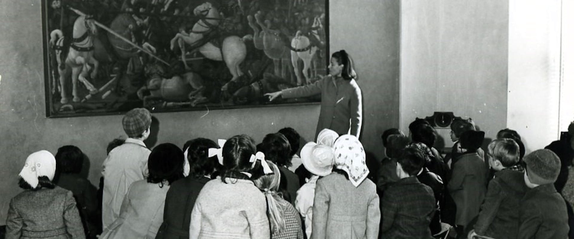 Maria Fossi Todorov, fondatrice Sezione Didattica Uffizi Un gruppo di bambini in abiti invernali è seduto su sgabelli pieghevoli, mentre una guida indica un grande dipinto storico all'interno di una galleria d'arte.