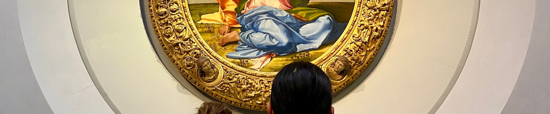 Two people stand closely together, admiring a circular Renaissance painting adorned with a detailed, ornate golden frame in a well-lit gallery setting.