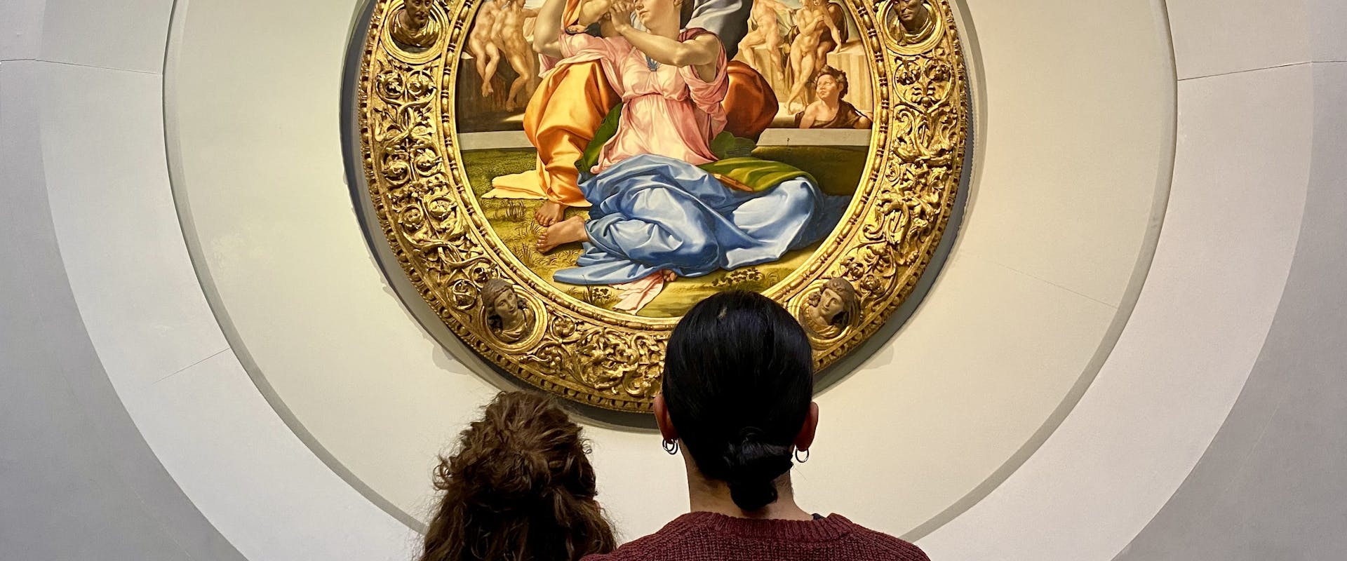 Two people stand closely together, admiring a circular Renaissance painting adorned with a detailed, ornate golden frame in a well-lit gallery setting.