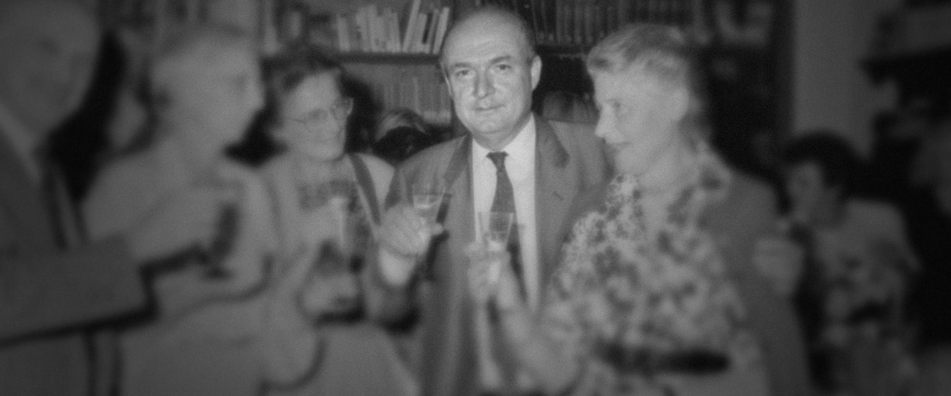 In una sala affollata, un gruppo di persone eleganti brinda con bicchieri di champagne davanti a uno sfondo di scaffali pieni di libri.