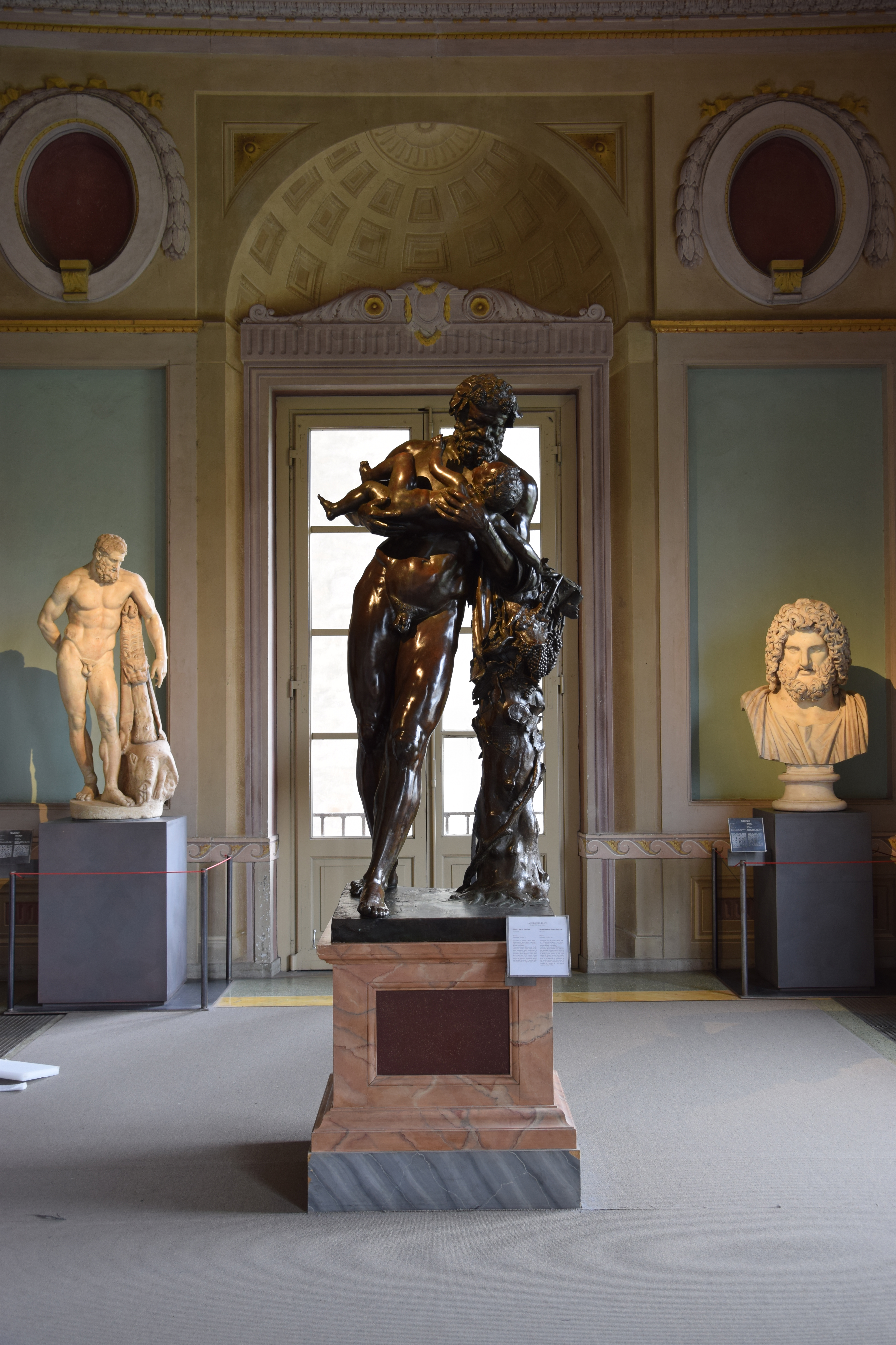 A classical museum interior showcasing a central bronze sculpture surrounded by other marble statues, with a softly lit detailed ceiling and intricate architectural elements.