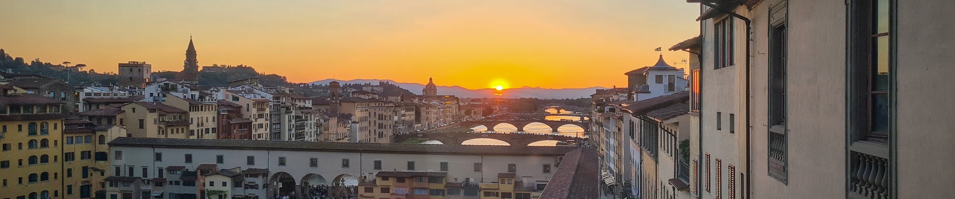 A breathtaking view of the Florence skyline at sunset highlights the iconic Ponte Vecchio bridge spanning the serene Arno River, with vibrant hues of orange and gold illuminating the historic architecture.