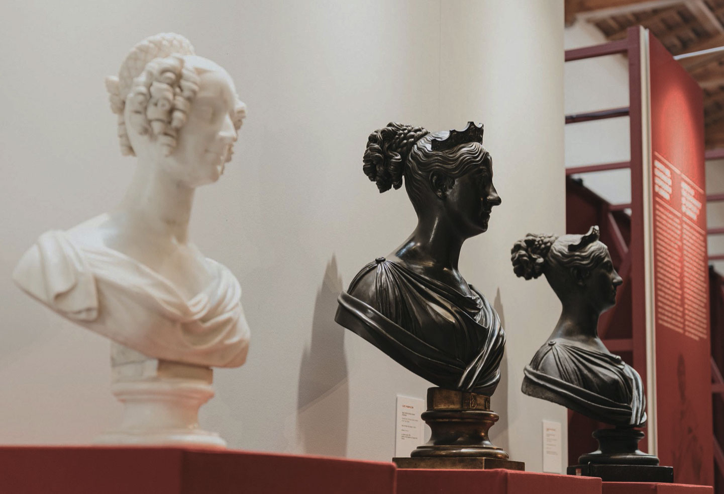 Tre busti in marmo e bronzo su piedistalli rossi in mostra in un museo, con uno sfondo di pannelli informativi.