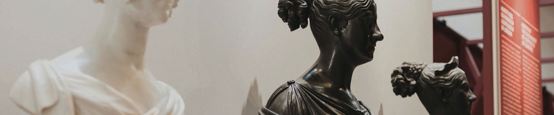 Tre busti in marmo e bronzo su piedistalli rossi in mostra in un museo, con uno sfondo di pannelli informativi.