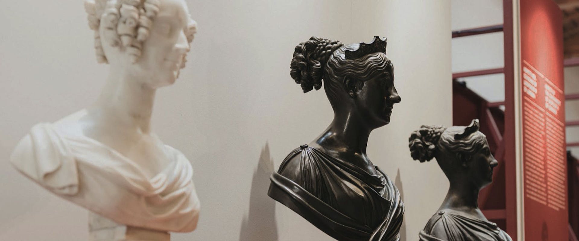 Tre busti in marmo e bronzo su piedistalli rossi in mostra in un museo, con uno sfondo di pannelli informativi.