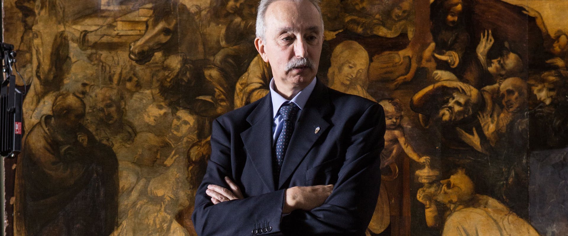 Un uomo con baffi in abito scuro è in piedi davanti a un complesso affresco antico che raffigura una scena religiosa dettagliata, donando un aspetto di autorità e contemplazione.