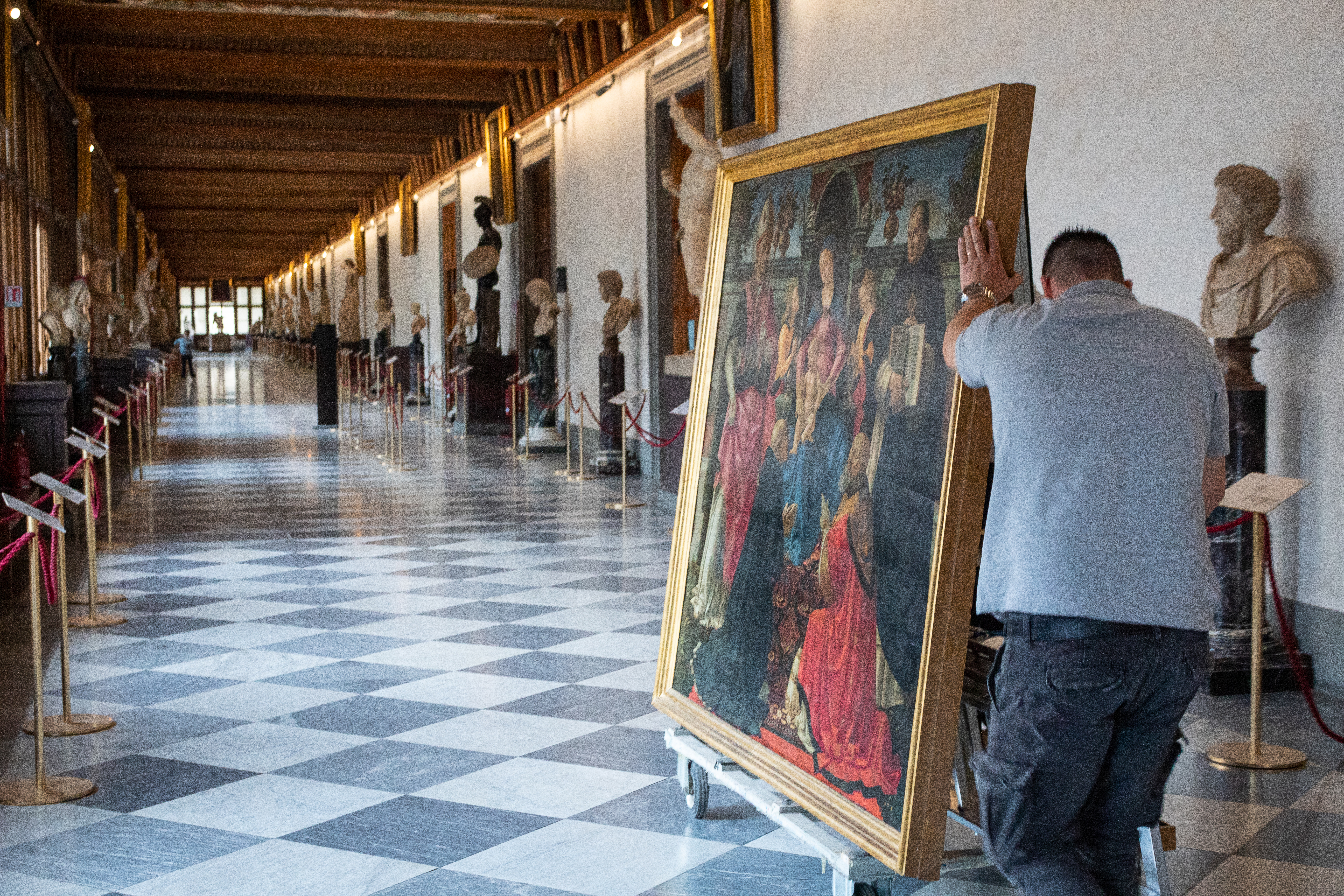 Un uomo sposta un grande quadro lungo un corridoio spazioso adornato con statue e busti in marmo, pavimento a scacchi in bianco e nero sotto un soffitto decorativo in legno.