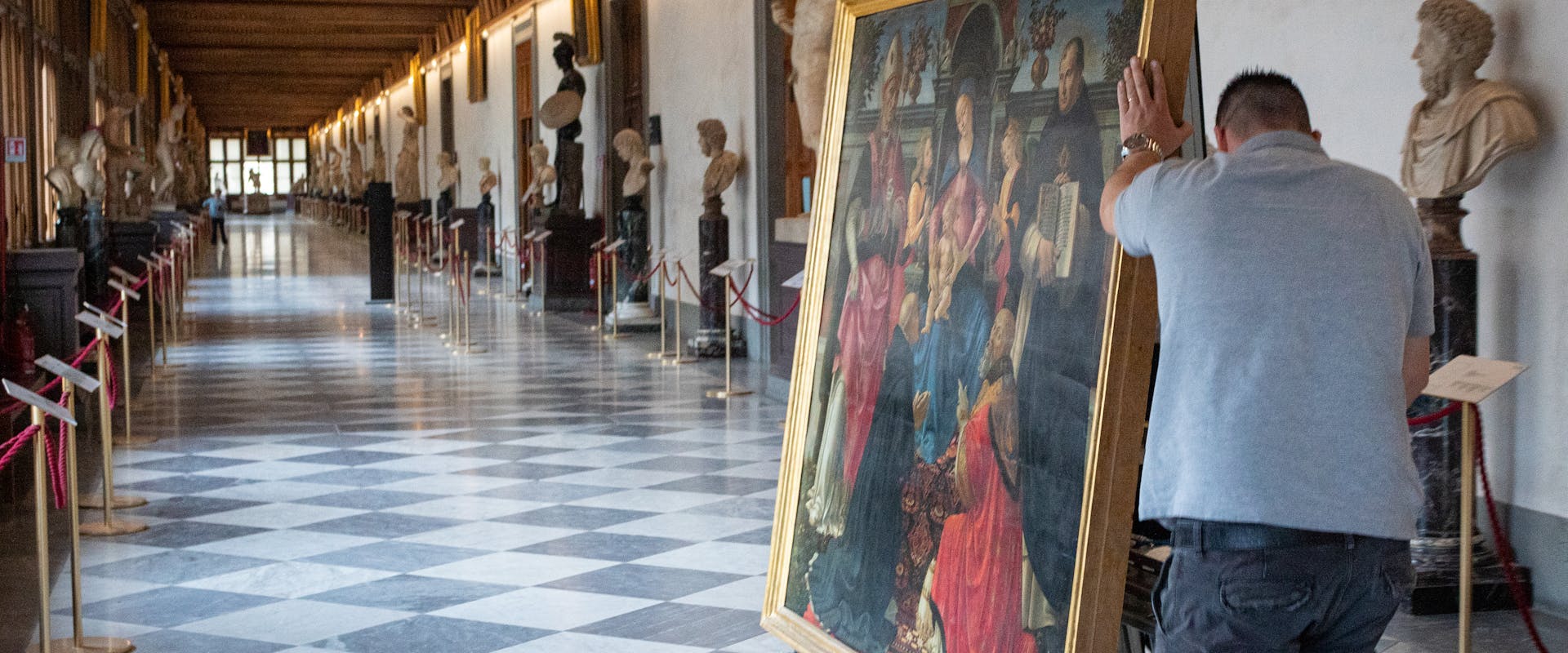 A man pushes a large, framed painting on a wheeled cart through a museum corridor with checkered floors, surrounded by sculptures and classical busts displayed along the walls.
