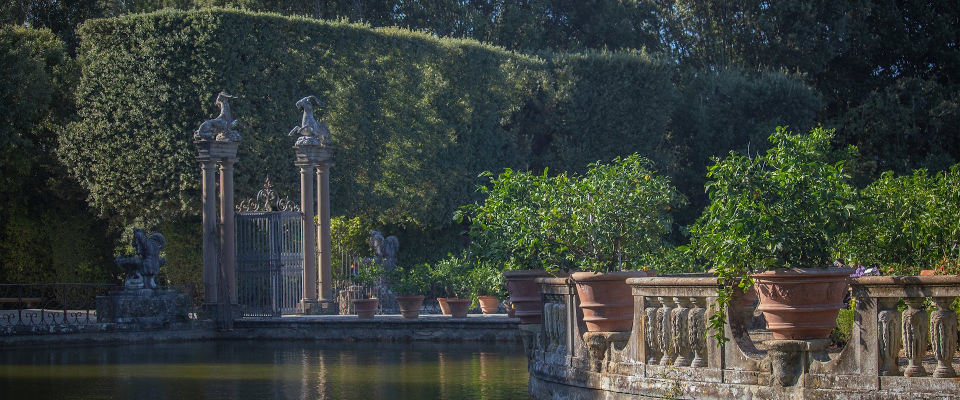 Giardino sontuoso con un ampio stagno circondato da vasi di terracotta e piante rigogliose, delimitato da intricati cancelli di ferro battuto e siepi curate sotto un cielo blu limpido.