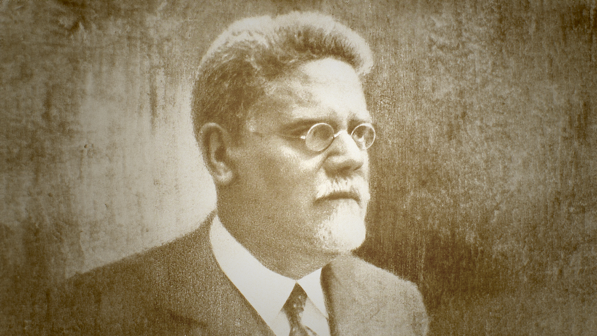 Una foto in bianco e nero di un uomo anziano con barba e occhiali rotondi, vestito in modo formale con un abito scuro, su uno sfondo sfocato e monocromatico.