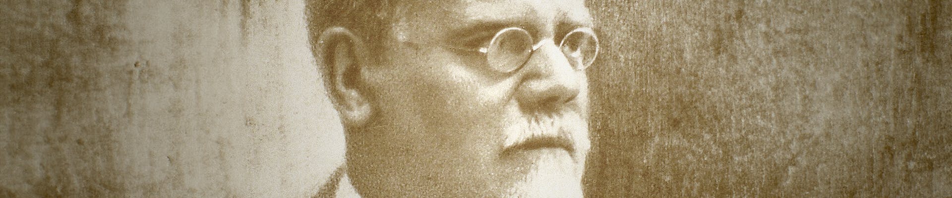 Una foto in bianco e nero di un uomo anziano con barba e occhiali rotondi, vestito in modo formale con un abito scuro, su uno sfondo sfocato e monocromatico.