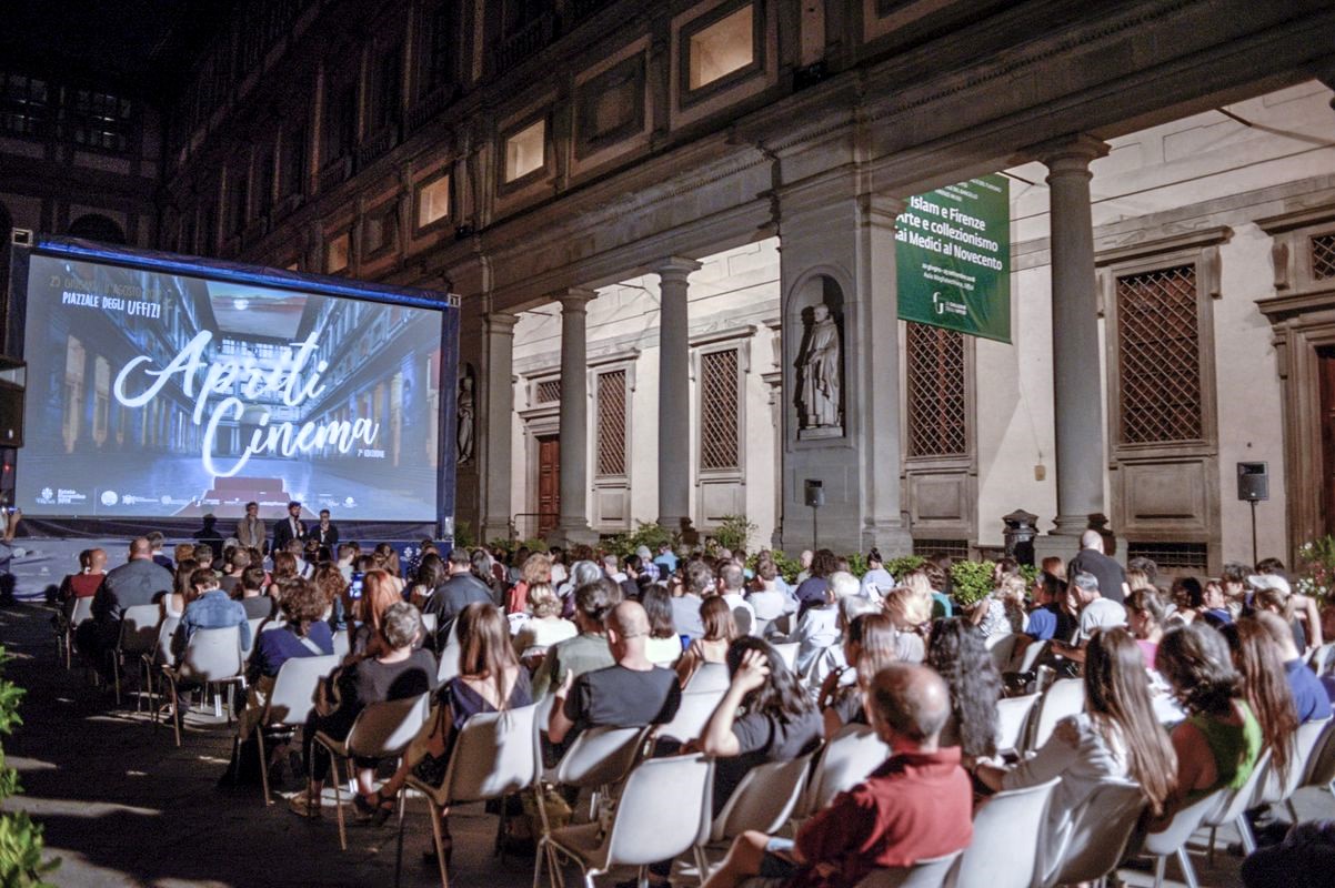 Persone sedute in un cortile notturno, davanti a uno schermo gigante che proietta un film sotto un cielo sereno durante un evento di cinema all'aperto.