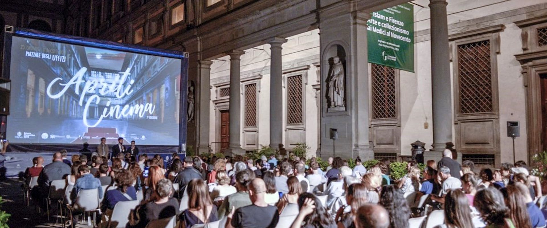 Persone sedute in un cortile notturno, davanti a uno schermo gigante che proietta un film sotto un cielo sereno durante un evento di cinema all'aperto.