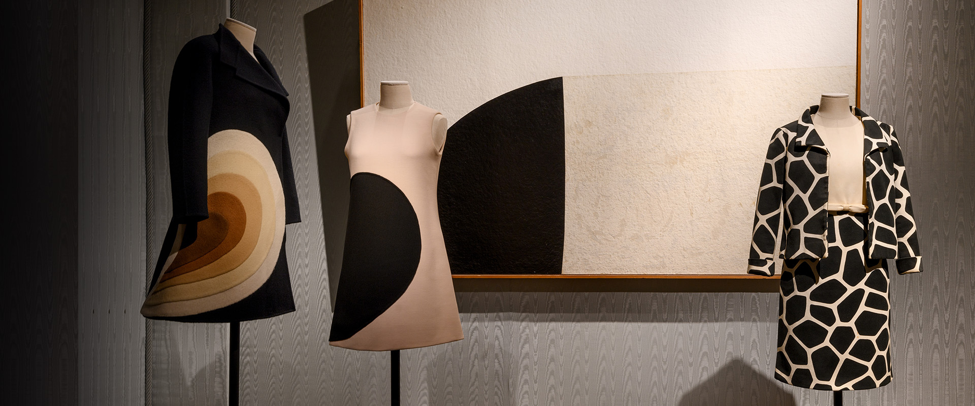 Tre manichini esibiscono abiti vintage con design geometrici audaci di diverse tonalità di nero, bianco e marrone, posizionati davanti a un grande quadro astratto che riflette motivi simili.