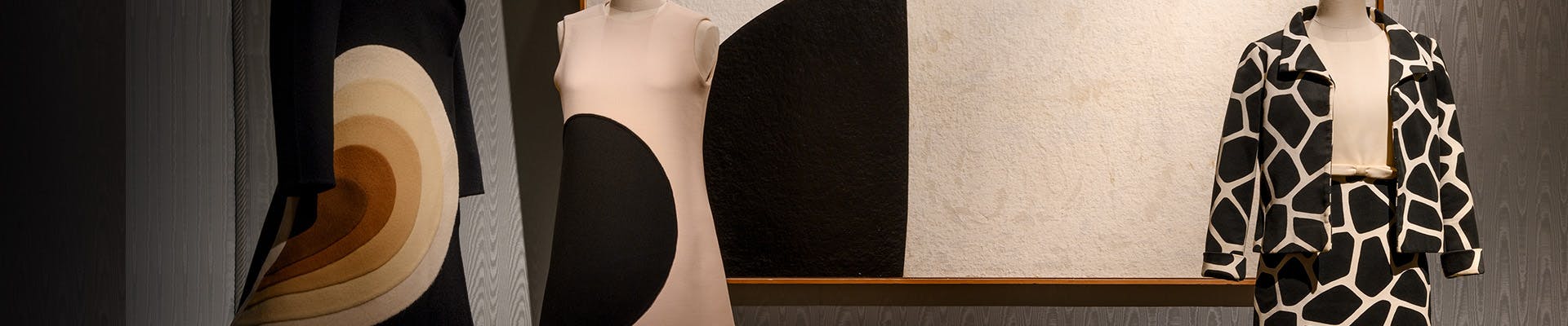 Il Museo della Moda e del Costume Tre manichini esibiscono abiti vintage con design geometrici audaci di diverse tonalità di nero, bianco e marrone, posizionati davanti a un grande quadro astratto che riflette motivi simili.