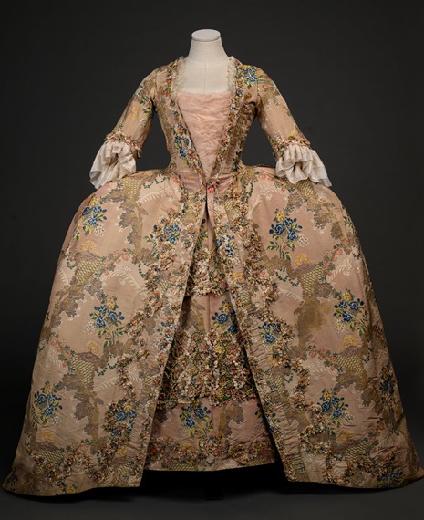 Abito storico del XVIII secolo in tessuto floreale rosa e oro, caratterizzato da una gonna ampia e maniche ornate con volant bianchi, perfetto esempio di moda aristocratica dell'epoca.