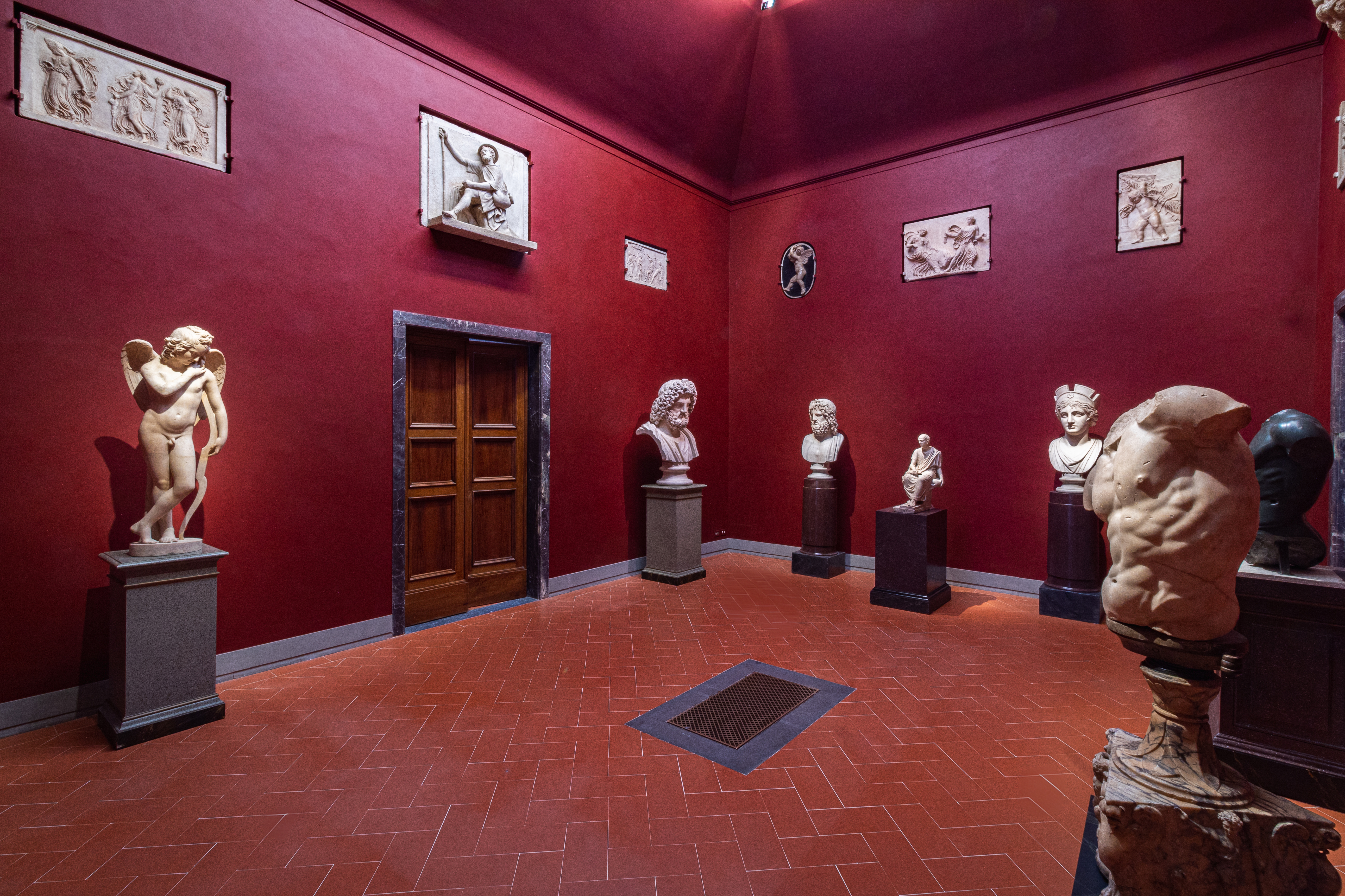 Una stanza di un museo d'arte con pareti rosse accoglie diverse sculture classiche in marmo disposte su piedistalli, illuminata da luci soffuse provenienti dal soffitto alto.