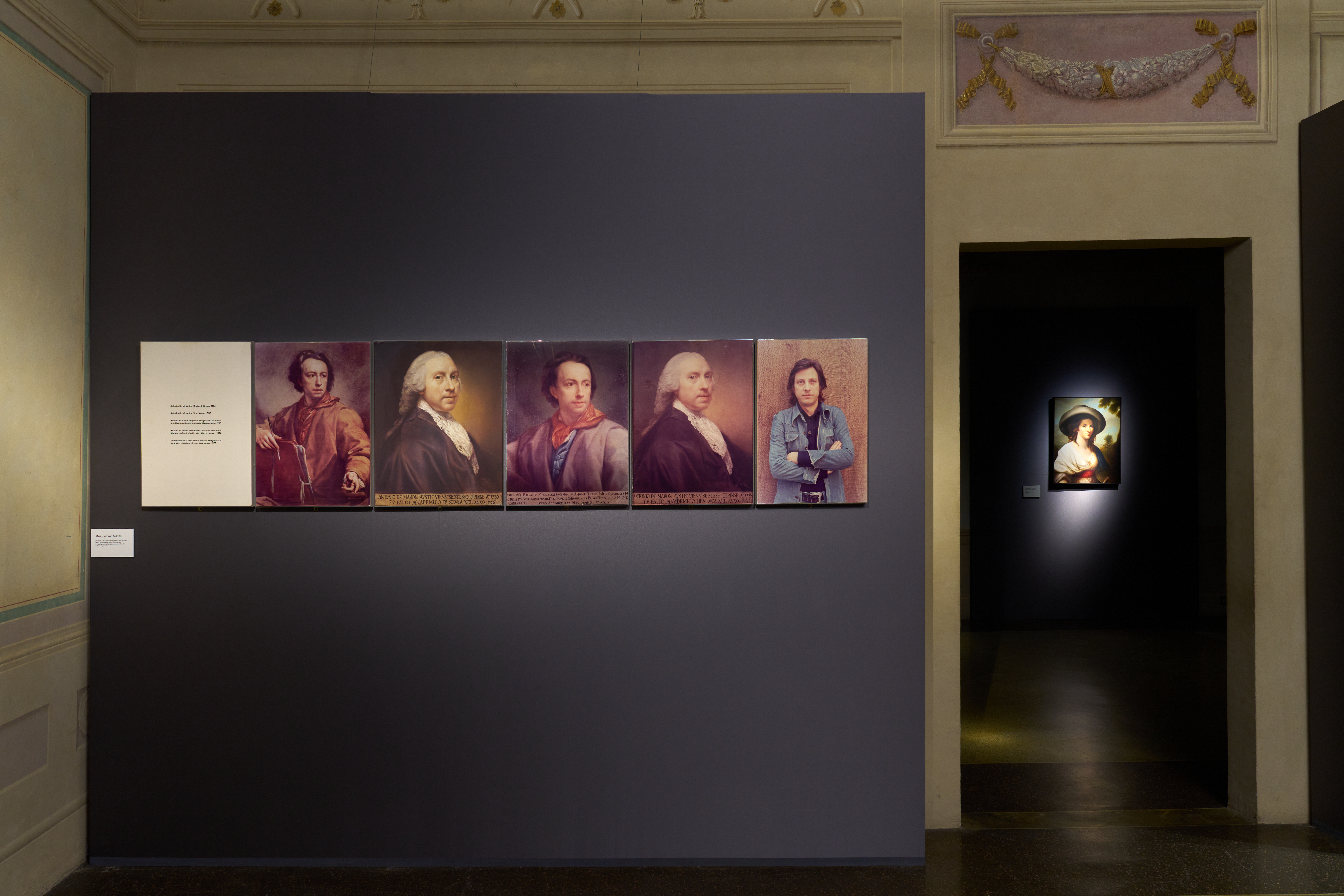 A gallery exhibit showcasing five historical portrait paintings on a dark gray wall, each depicting different individuals with varied attire and expressions, alongside a smaller illuminated portrait in an adjacent dimly lit room; the scene conveys a strong sense of tradition and artistic heritage.