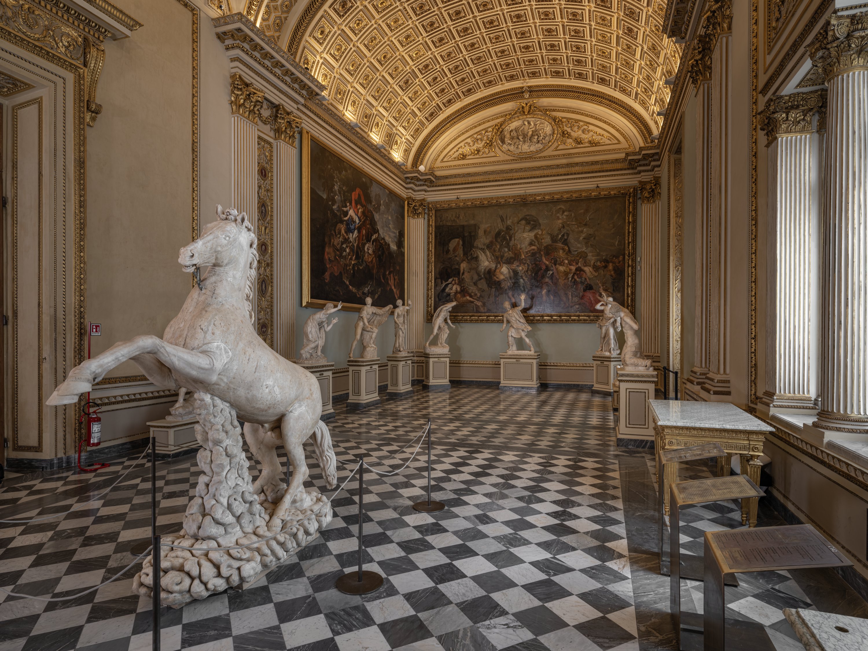 Un'elegante sala di un museo con statue di marmo, tra cui un cavallo bianchissimo in primo piano, un pavimento a scacchiera bianco e nero e soffitti riccamente decorati, illuminata da luce naturale morbida, con dipinti storici appesi alle pareti.