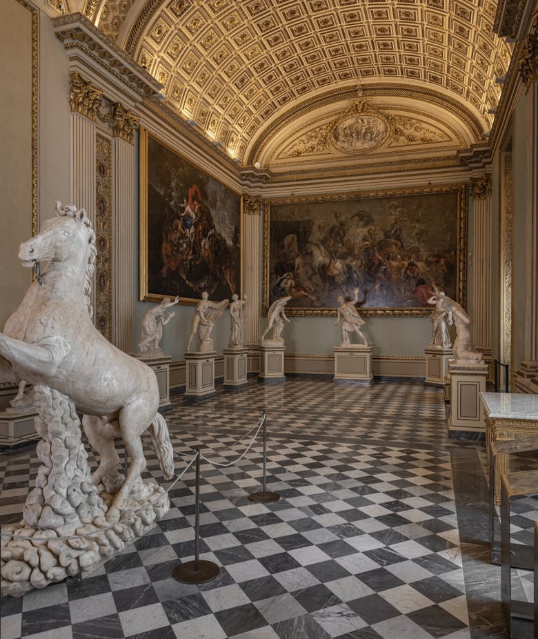 An elegant gallery with a grand arched ceiling showcases a series of white marble statues, including a prominent horse sculpture, all set on a checkered black-and-white marble floor, enhancing the room's opulent ambiance.