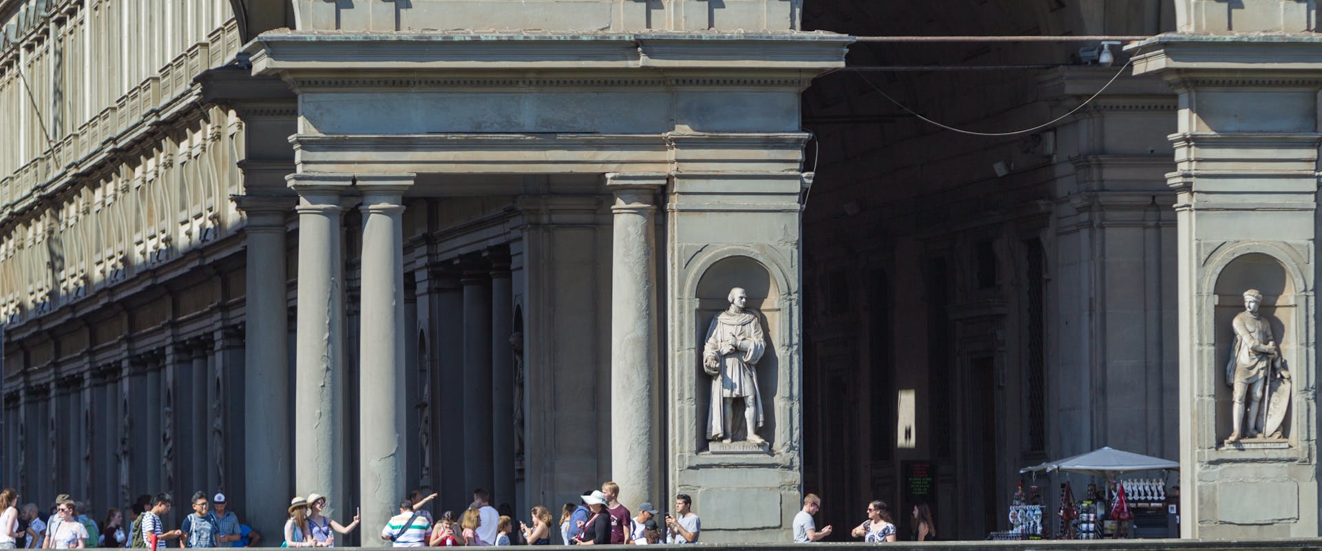 Il corridoio degli Uffizi a Firenze, con i suoi archi classici, colonne imponenti e numerose statue, affollato di visitatori che camminano e si fermano per ammirare l'architettura storica.