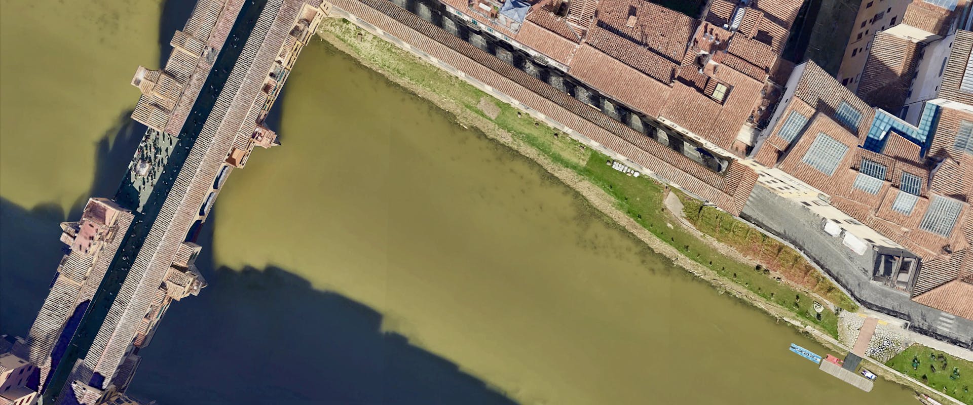 Veduta aerea di una sezione del fiume con l'antico ponte di pietra, edifici storici con tetti rossi ai lati, e un'area verde lungo la riva.