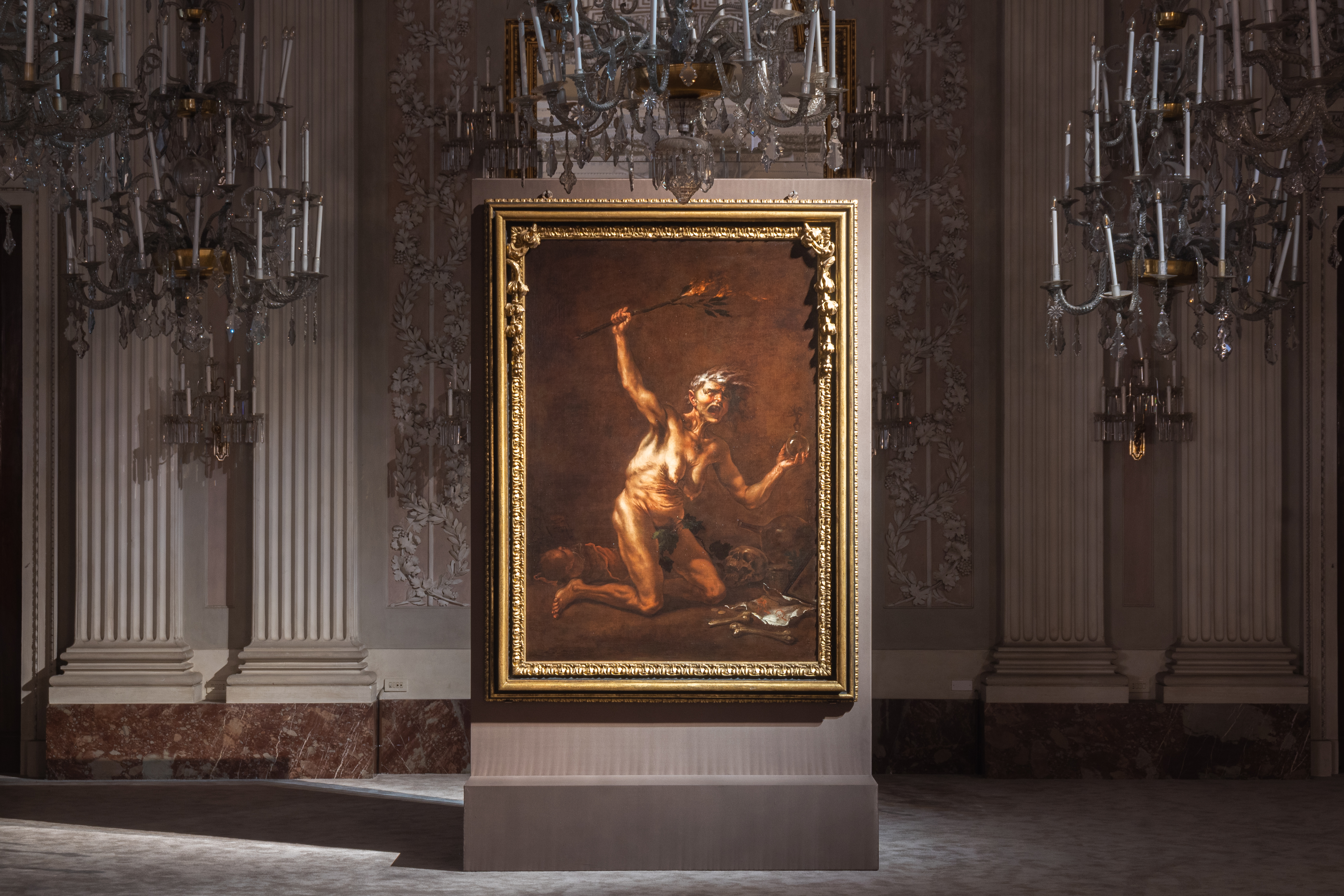A classic painting with a gilded frame is displayed in an opulent room adorned with ornate chandeliers and intricate wall details, highlighting the artwork's historic and artistic significance.