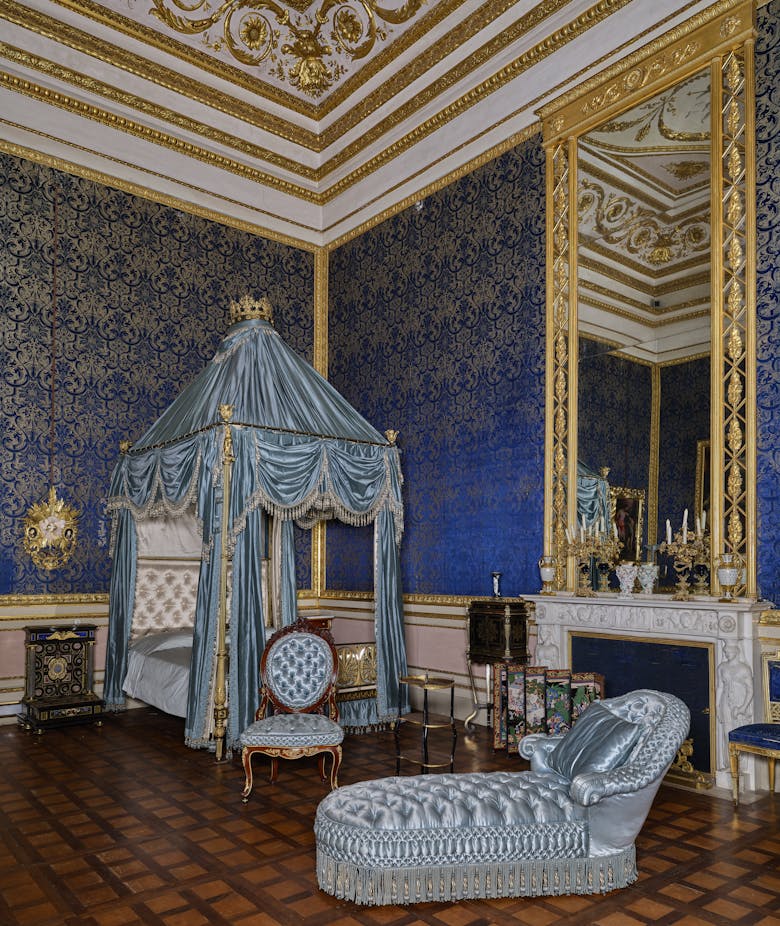 Luxurious blue and gold royal bedroom with ornate canopy bed, elegant chaise lounge, and intricate wall patterns, highlighting opulent interior design and classic architecture.