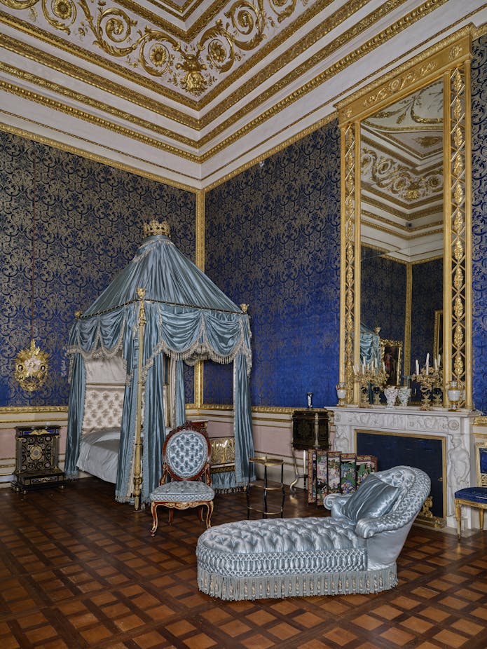 Una lussuosa stanza con pareti blu decorate, un soffitto ornato d'oro, un elegante baldacchino blu con tende drappeggiate, e un divano chaise longue abbinato.