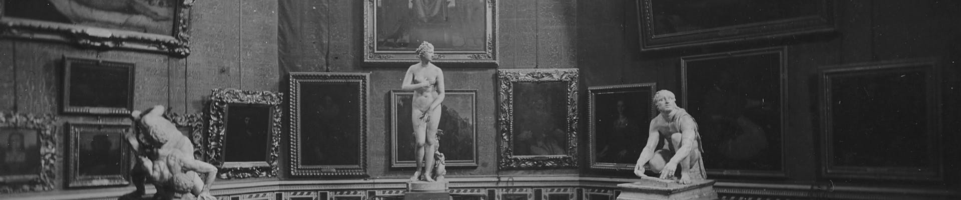 Una galleria d'arte con diverse opere appese alle pareti, tra cui dipinti incorniciati e due sculture classiche su piedistalli, illuminata dalla luce naturale che entra dall'alto.