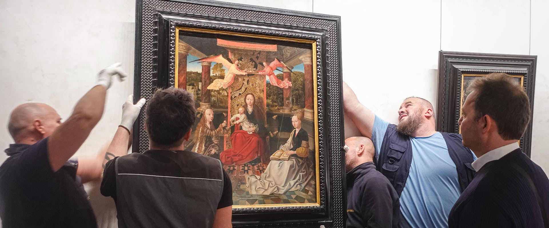 Maestro di Hoogstraeten, Madonna con Bambino in trono con Santa Caterina d'Alessandria e Santa Barbara, 1500-10 c., Uffizi Cinque persone collaborano per appendere un grande dipinto in una cornice nera su una parete chiara, in una galleria d'arte.