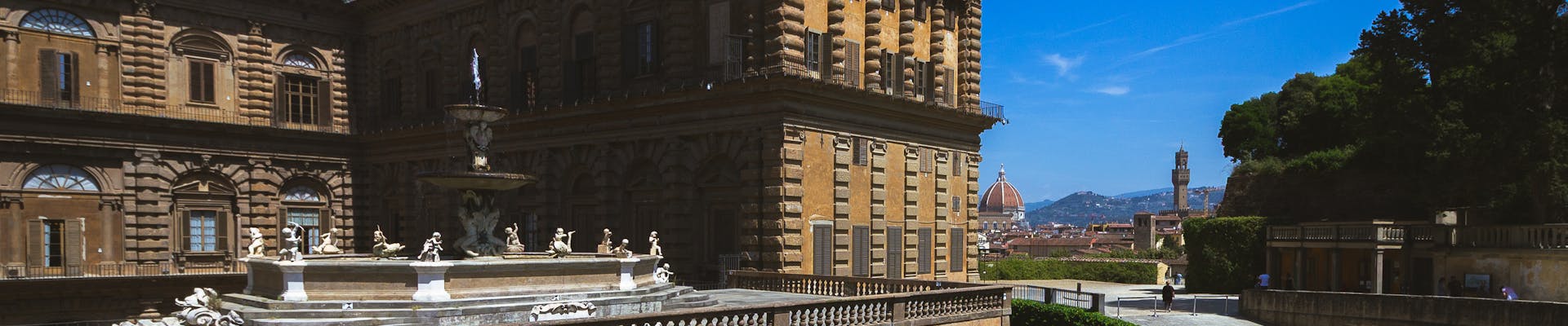 L'immagine mostra il Palazzo Pitti a Firenze, con i suoi giardini ben curati e la magnifica architettura rinascimentale, sotto un cielo azzurro e luminoso, con la cupola del Duomo visibile in lontananza.