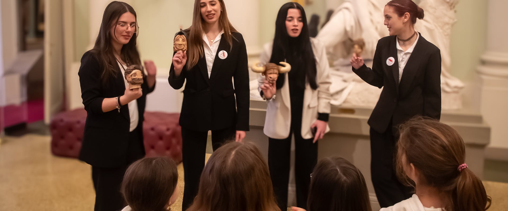 Un gruppo di donne in abiti formali tiene delle statuette mentre parla a bambini seduti in un elegante museo con una statua classica sullo sfondo.