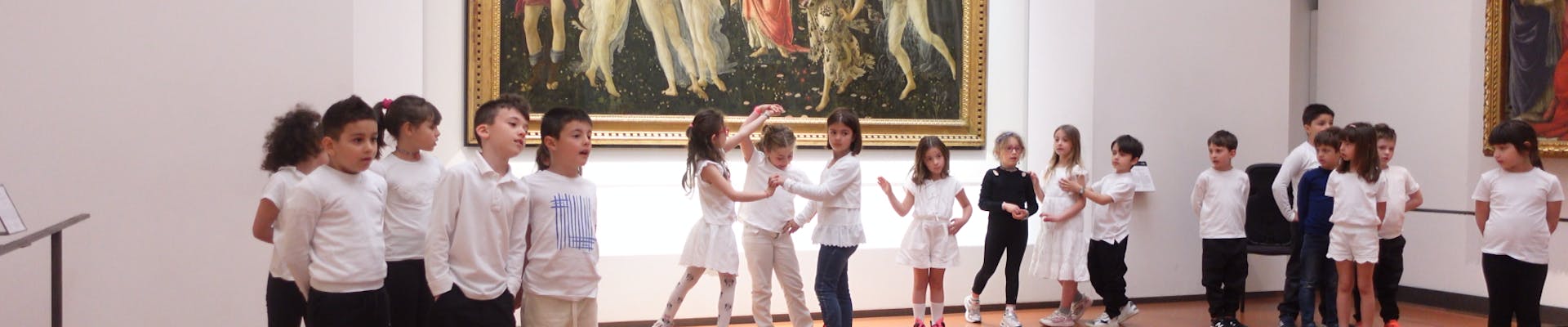 Nel museo, un gruppo di bambini in magliette bianche e pantaloni scuri è disposto in due file di fronte a un grande dipinto rinascimentale appeso alla parete, creando un contrasto tra arte classica e presenza moderna.