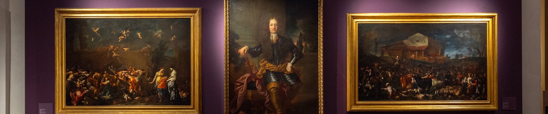 Tre dipinti in cornici dorate sono appesi su una parete viola in una galleria d'arte, con un pavimento in mattoni che contribuisce all'atmosfera classica ed elegante.