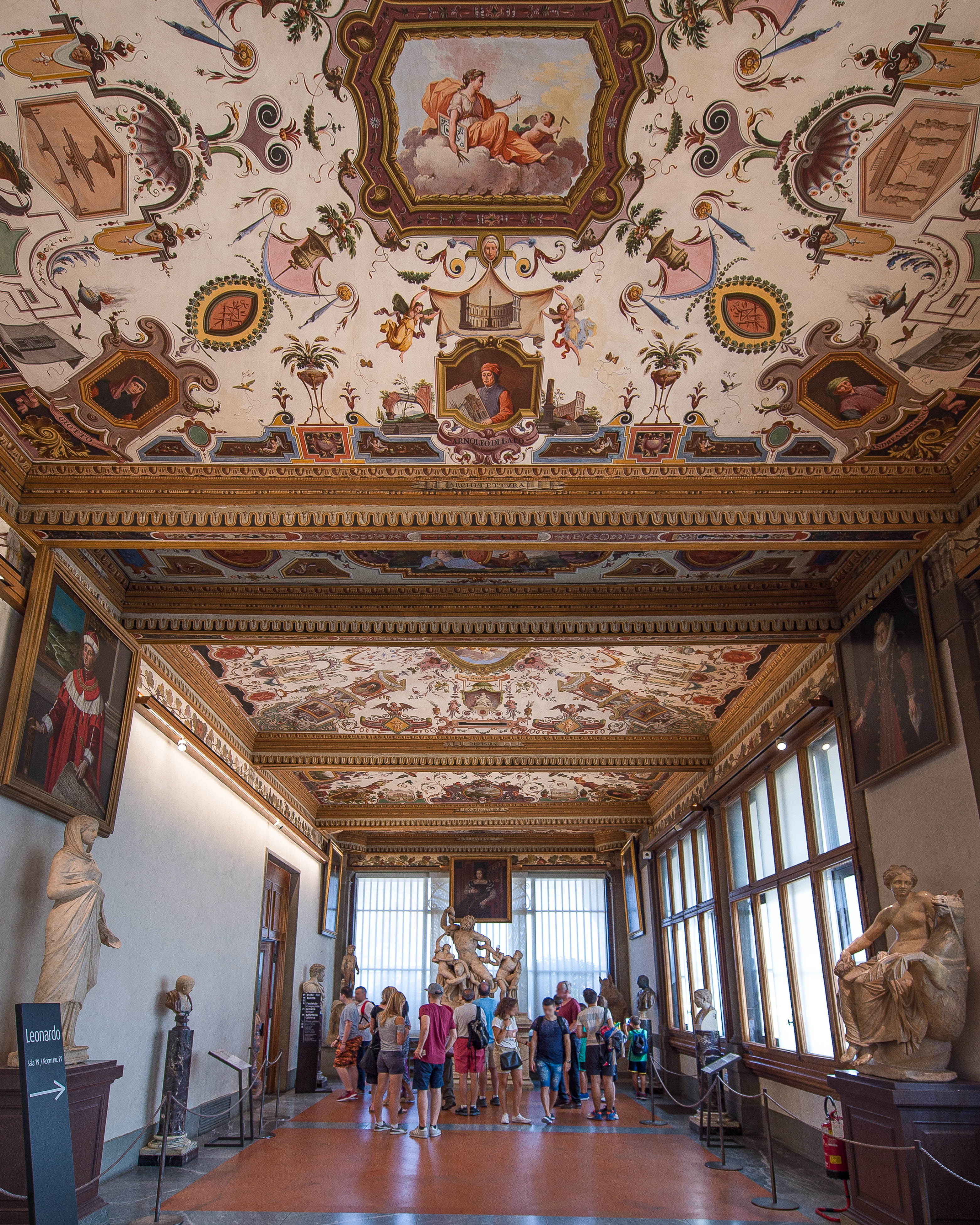 Il soffitto decorato di una galleria d'arte storica presenta intricati motivi floreali e affreschi, mentre sculture di marmo e dipinti ornano le pareti, con visitatori che ammirano le opere.