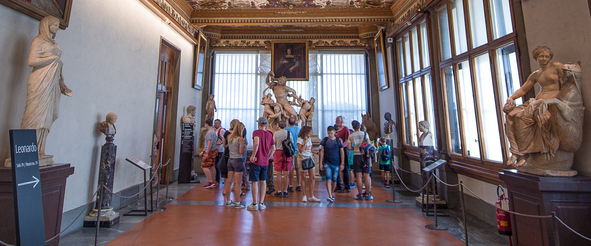 Il soffitto decorato di una galleria d'arte storica presenta intricati motivi floreali e affreschi, mentre sculture di marmo e dipinti ornano le pareti, con visitatori che ammirano le opere.