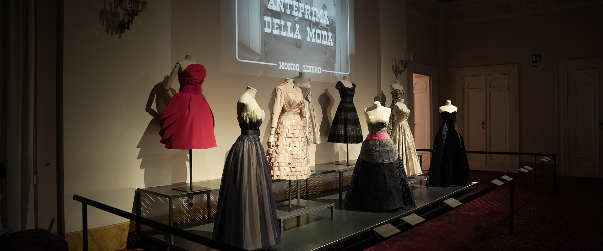In mostra, una fila di eleganti abiti da sera su manichini, con un abito rosso acceso in contrasto con una serie di vestiti nei toni del beige e nero, in una sala espositiva dall'illuminazione soffusa; l'arredamento include uno schermo con titoli di testo sfocati dietro la scena.