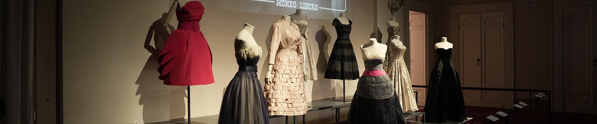 In mostra, una fila di eleganti abiti da sera su manichini, con un abito rosso acceso in contrasto con una serie di vestiti nei toni del beige e nero, in una sala espositiva dall'illuminazione soffusa; l'arredamento include uno schermo con titoli di testo sfocati dietro la scena.