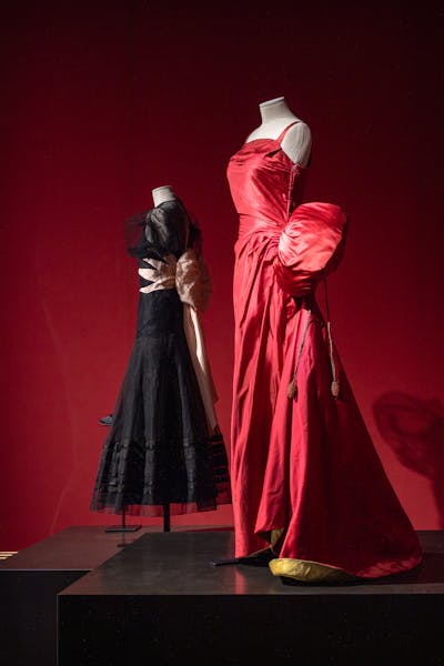 Due manichini esposti su sfondo rosso, uno indossa un abito nero con dettagli floreali bianchi in vita, l'altro è vestito con un abito rosso luminoso con maniche voluminose, entrambi rappresentano un'elegante moda da sera.