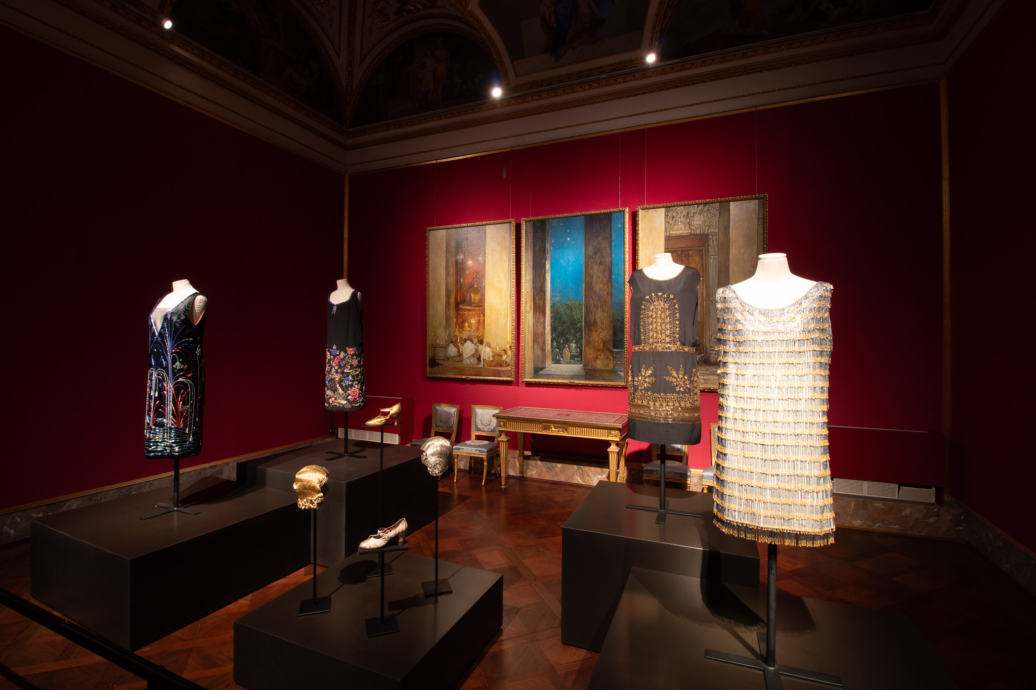 Una mostra d'arte in un museo con abiti eleganti su manichini, insieme a maschere decorative, esposti su piedistalli scuri in una stanza dalle pareti rosse con un grande dipinto appeso sullo sfondo.