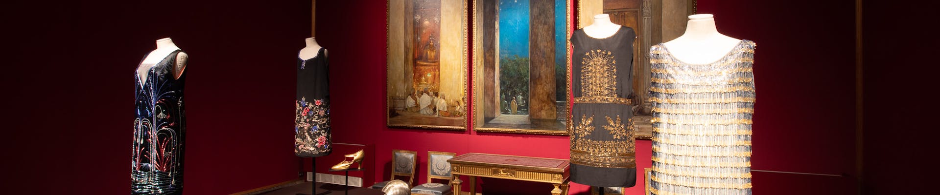 Una mostra d'arte in un museo con abiti eleganti su manichini, insieme a maschere decorative, esposti su piedistalli scuri in una stanza dalle pareti rosse con un grande dipinto appeso sullo sfondo.