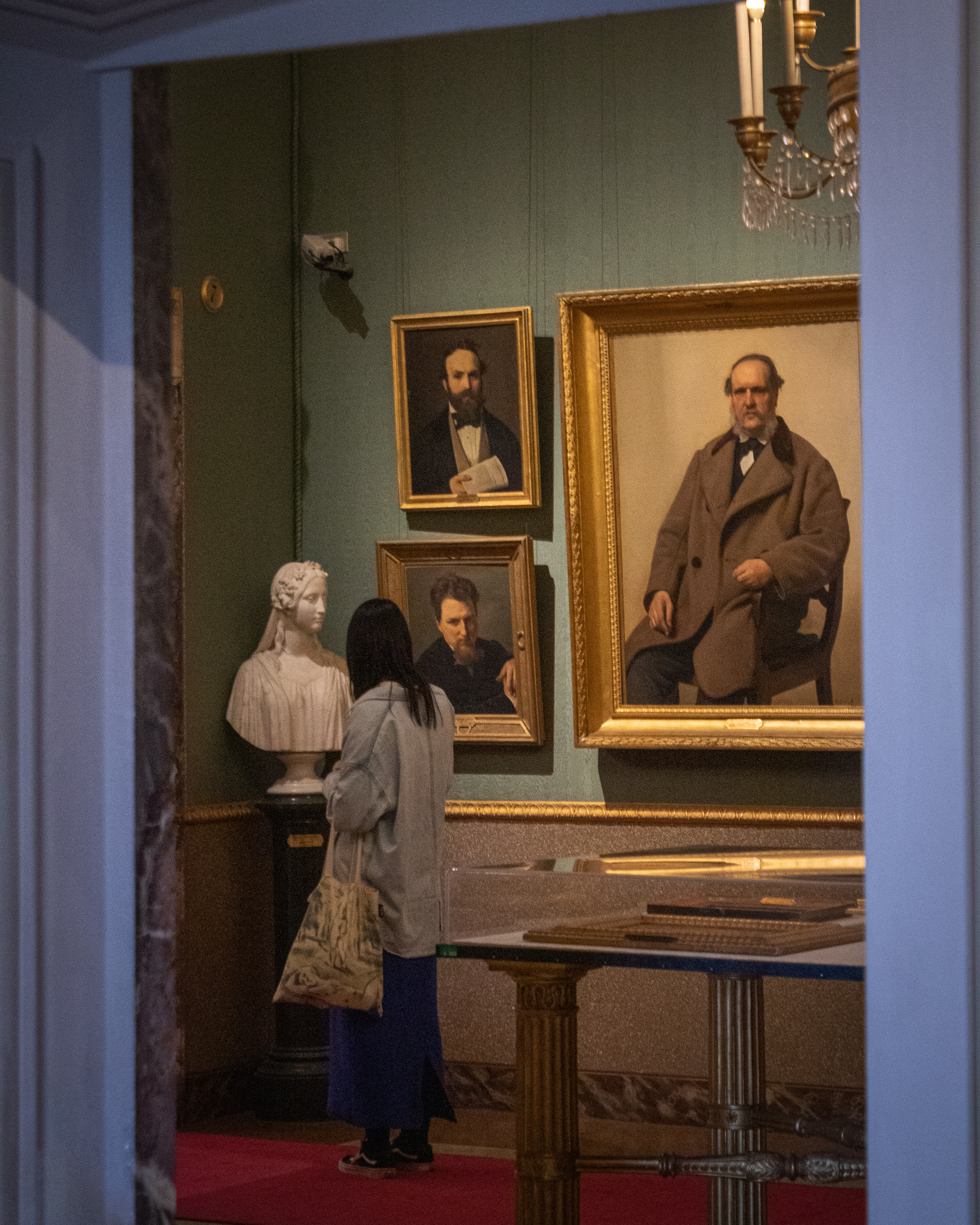 Una persona osserva ritratti e un busto in una sala di un museo, con pareti verdi e un tavolo decorativo al centro, illuminati da un lampadario elegante.