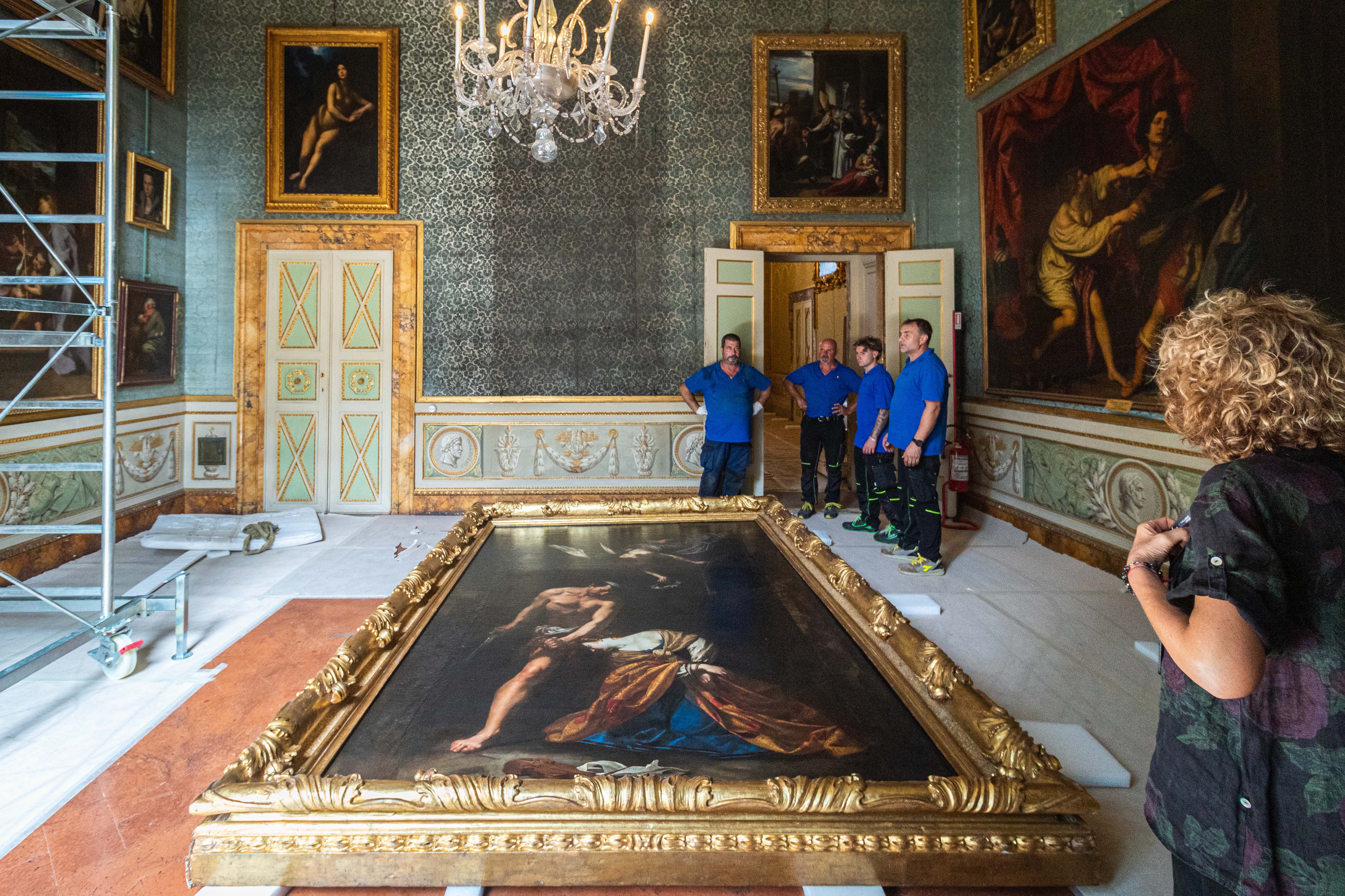 A large, ornately framed painting lies on the floor of an opulent room with high ceilings, adorned with classical art and a grand chandelier, as several people in blue shirts stand nearby, possibly preparing for its installation or restoration.