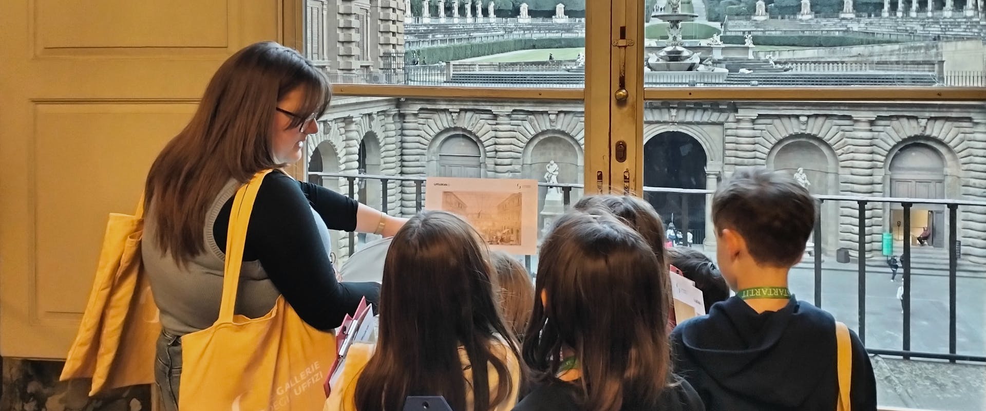 Un gruppo di bambini ascolta attentamente una donna che spiega davanti a una finestra che si affaccia su un museo storico, con dettagli architettonici imponenti visibili sullo sfondo.