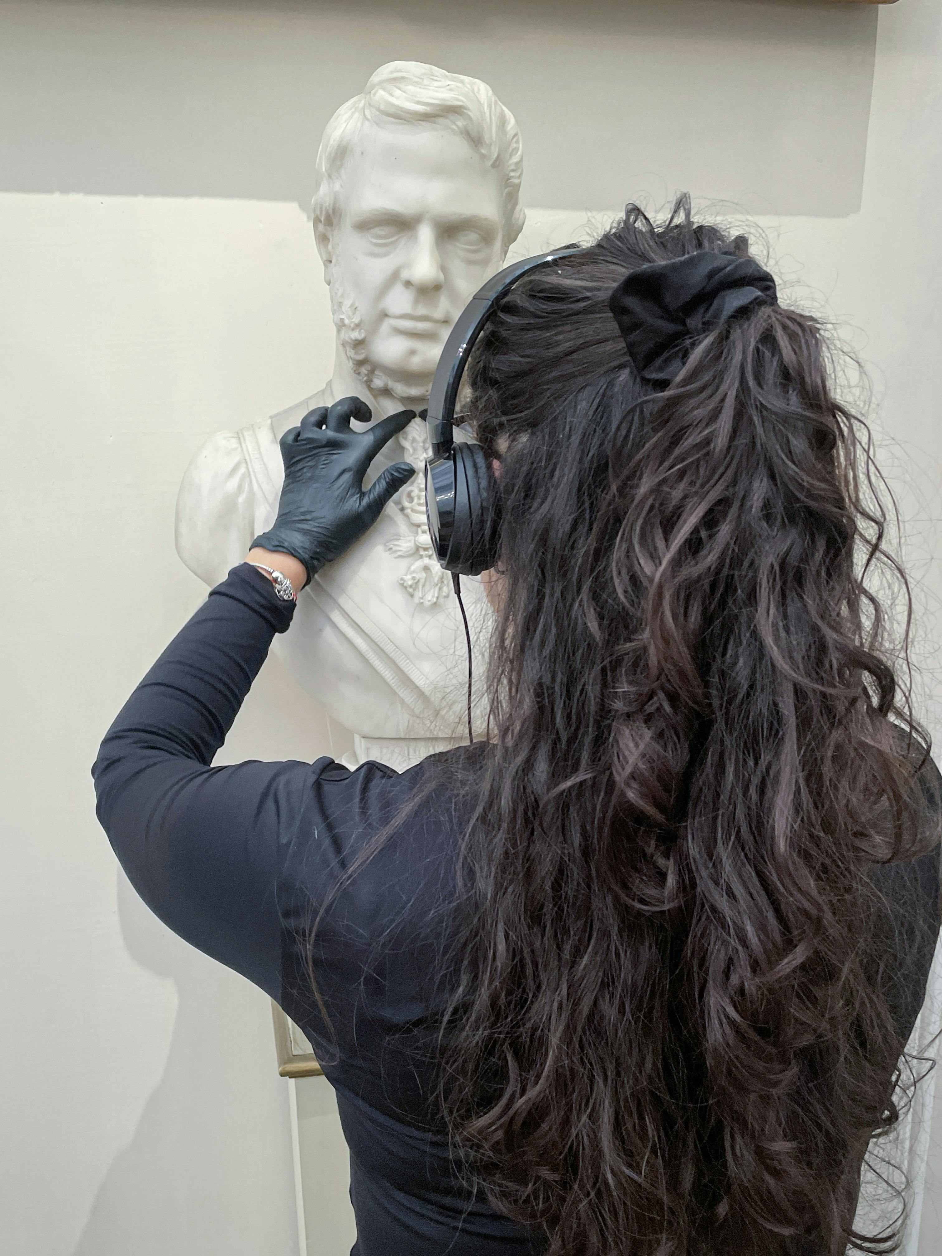 A person with long dark hair, wearing black clothing and gloves, listens to a white marble bust using headphones in a museum setting.