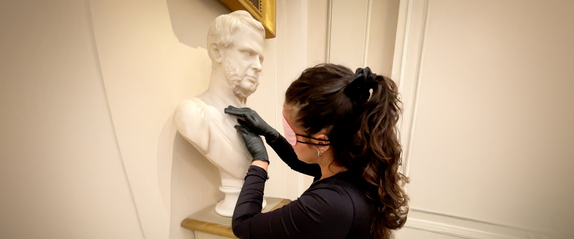 A woman with long, dark hair and black gloves carefully cleans a white marble bust displayed on a wooden pedestal in an elegantly decorated room, indicating an art restoration process in a museum or gallery setting.