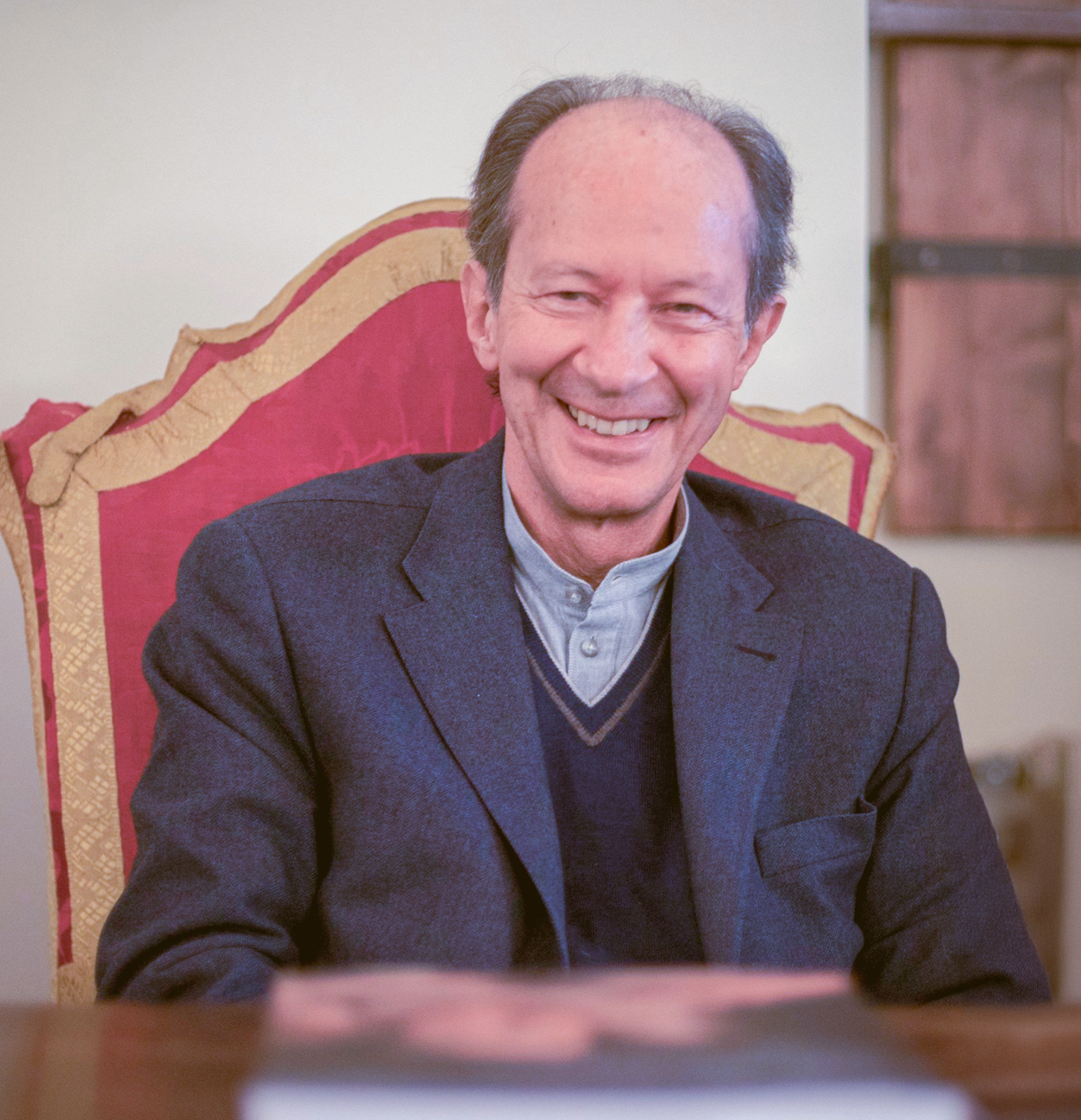 Giorgio Agamben siede sorridente su una sedia ornata, indossando una giacca elegante sopra una camicia chiara e un maglione scuro, con un libro sfocato in primo piano.